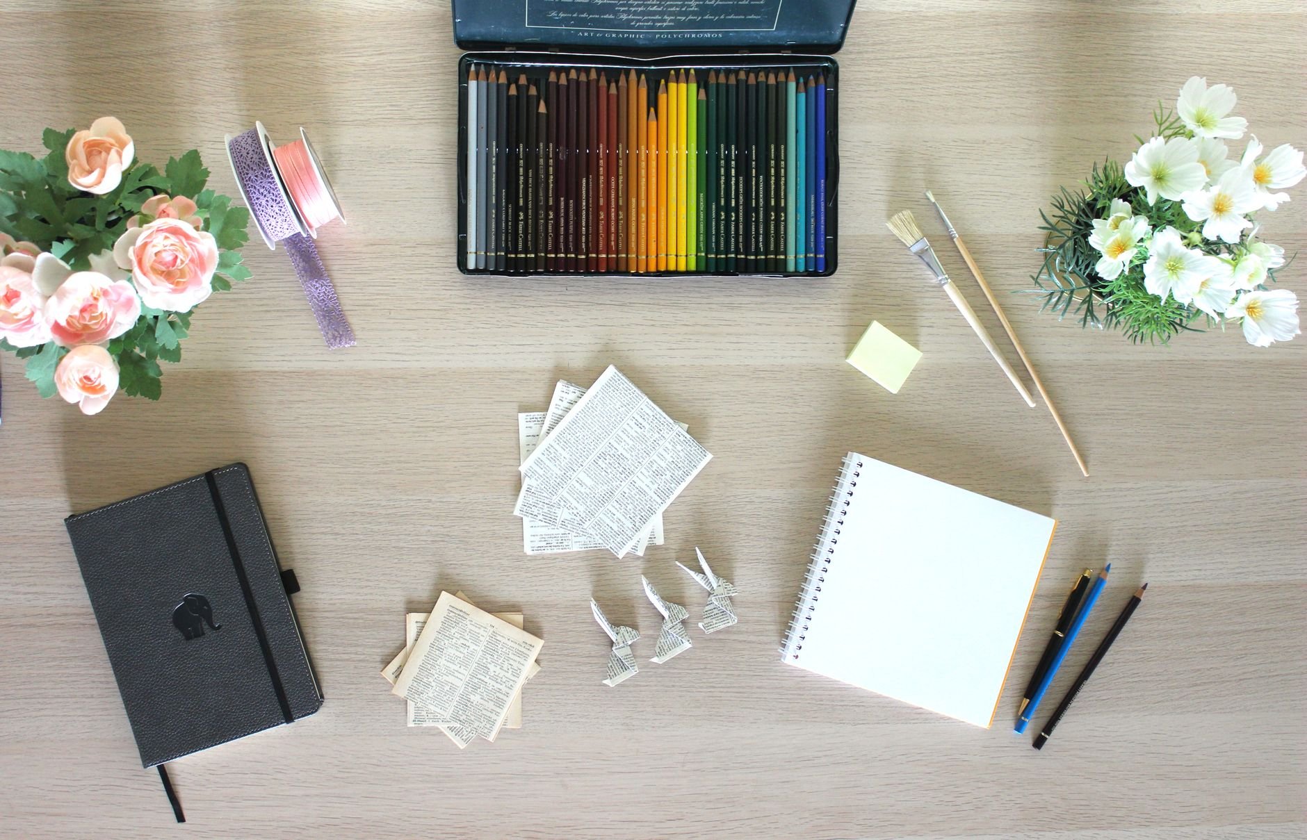 Flat lay of art supplies and stationery on a wooden table, including flowers.